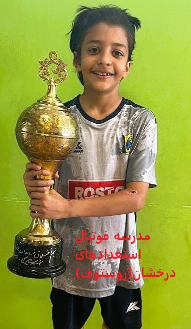 فوتبالیست نونهال با لباس طوسی و سورمه ای با لبخند و جام قهرمانی در دست